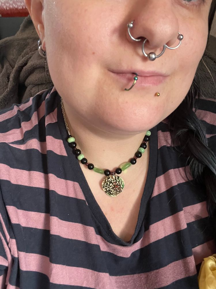 selfie of sinead shown from tip of nose - now featuring nostril rings on both sides with matching septum ring, down to top of striped shirt. fae is wearing a short necklace on a silver chain with black and jade green beads and a painted clay pendant which looks like mushrooms in greenery. very fae vibes. 