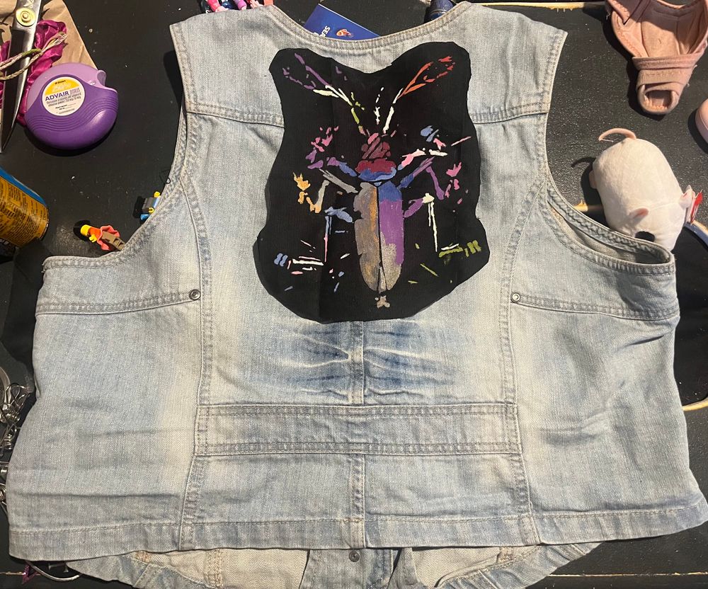 back of jacket on a table with various sundries. a black patch with dead channel sky “cricket” design spray-painted on it is at the top center, cutout vaguely to the contours of the bug, which has been painted in with various pride colors. 