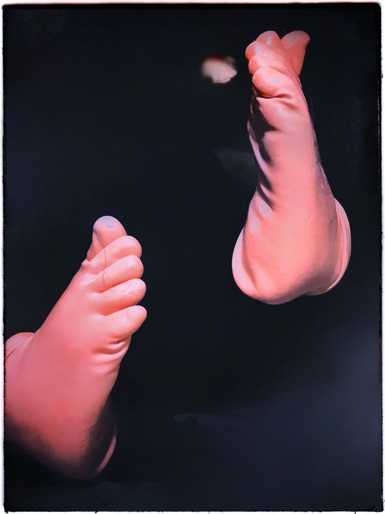 heavily edited image of latex sock-clad feet posed with soles facing each other and toes articulated. background has been faded to blurry shades of black and dark grey, except for mysterious glowing spot next to inside of left foot in upper right of frame. socks have an almost-fleshy light pink tone and crinkle around the bends and heels of the small feet. white frame added on image along with light glowy effect and slight hypersaturation elements. 