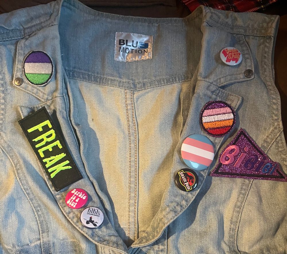 closeup of vest collar, showing from left to right circling the neckline (right side on left of frame): genderqueer circle glitter patch on top lapel, lower long lapel has black rectangle with “FREAK” in neon green, “barbie is a slut” pink pin with white bubble text, kiki’s delivery service pin. going up left lapel: jurassic park pin, trans pride larger pin, lesbian glitter pride flag patch (accidentally upside down, will be fixed) on bottom section with pink glitter triangle saying “BiBiBi” in bi pride colors to the side of other pride circles on lapel. top panel has light blue pin with “Gender is a Drag” in orange and red decorative text. 