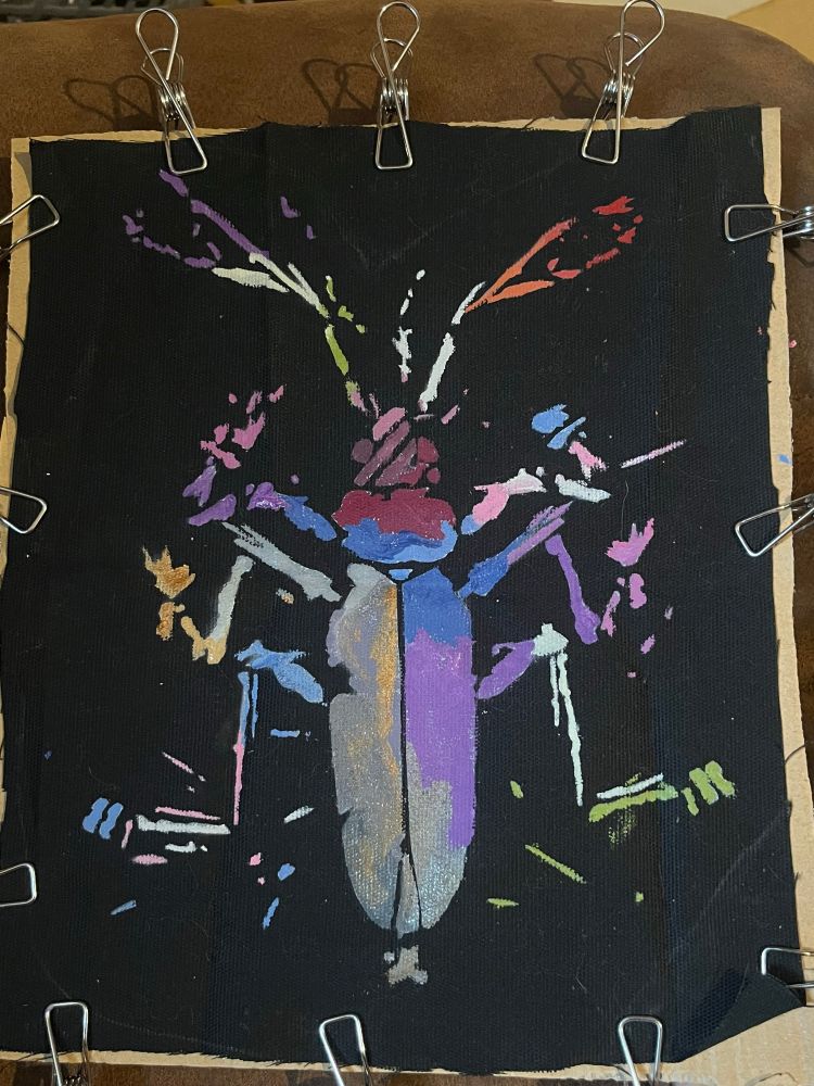 black patch drying clipped to cardboard before being cut out, showing color details. left antennae corner of bug is genderqueer pride, as is lowest right leg and part of body segment. top right antennae and head is lesbian 7-color pride flag, extending into part of upper body. upper left and middle right legs and body portions are bi pride, and bottom left and top right legs are trans pride. the lower left of body and middle left leg are shades of silver and grey with gold accents, more like an actual bug. 