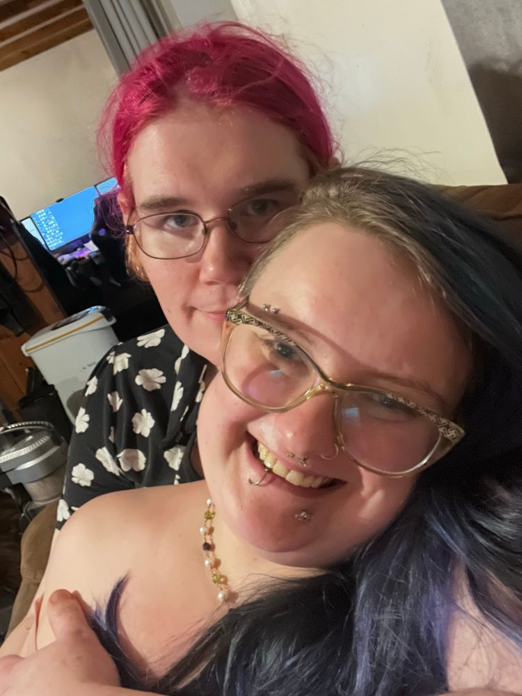 sinéad and lexi are seated on couch, lexi with pink hair pulled back, wire glasses on, and wearing a black dress with white floral print. she is holding sinéad seated in front of her, smiling softly with arm wrapped around the fae, who is wearing olive cat eye glasses and smiling wide through piercings, blurple hair obscuring clothing with bare shoulder out. 