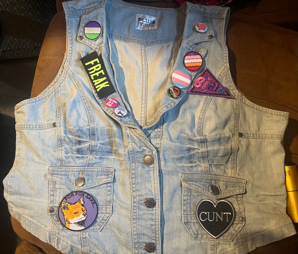 short light denim v neck vest layer out on couch. low pockets have patches laid on them, left is black heart saying “CUNT” in silver, and right is purple with orange fox and black text “For Fox Sake…”. collar is decked out in pins and patches, with triangle patch next to left lapel. 