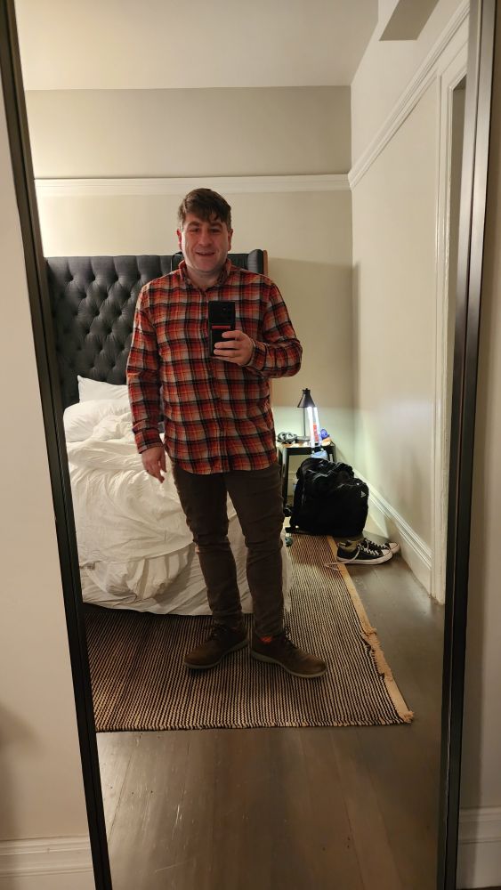 Full body shot in full length mirror. Orange and red plaid shirt with black check. Brown fitted pants. Brown rockports. Orange sock slightly showing.