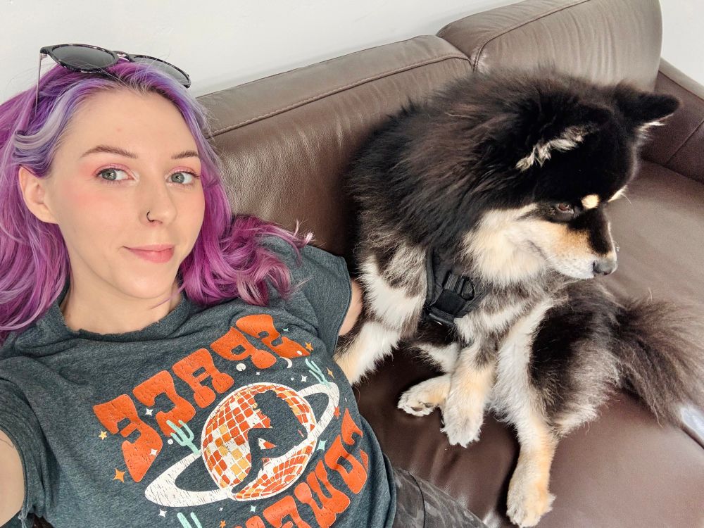 On a brown leather couch, I’m leaned back with my arm around my dog Bumble. My T-shirt says ‘space cowgirl’ in orange font. I have sunglasses on my head and my pinkish-purple hair is loosely curled. Bumble is sitting with his legs splayed to the side, looking away from the camera.