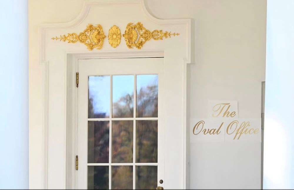 An entrance at the White House has tacky, fake gold crap above the door. Three papers are taped up that have gold cursive reading, 'The Oval Office.' The paper with the word, 'The,' is curling off the wall slightly.