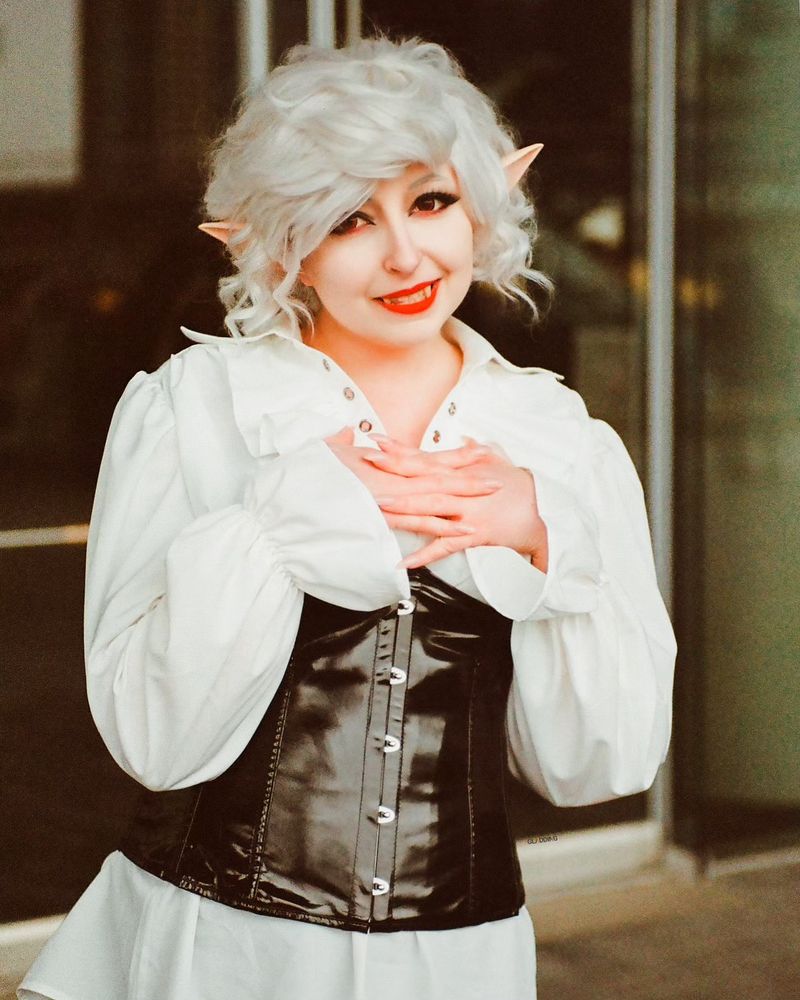 A femme Astarion cosplayer with hands crossed playfully over the chest, and a half smile. Wearing a flowy white blouse and a pleather corset. 