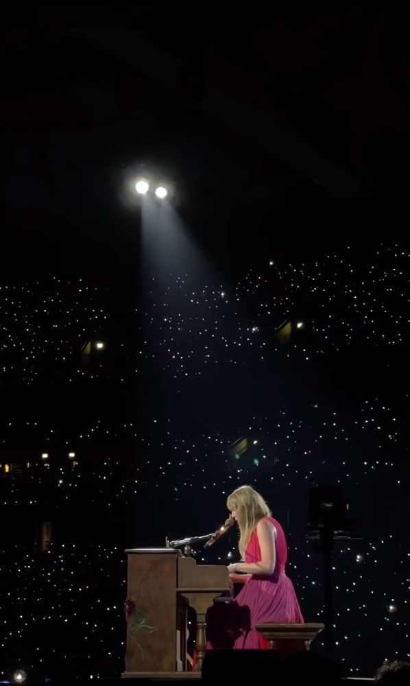 Taylor is sitting at her piano on the eras tour stage with all the twinkling light up wristbands in the background 