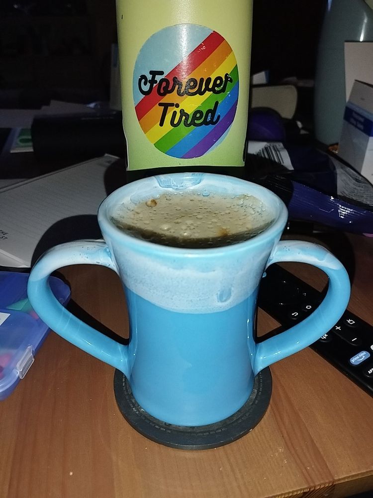 Blue cup with a handle on each side. Aka two handled cup. With a light blue/white top dipped rim. Background is a coffee table with a water bottle behind with a rainbow coloured sticker reading feeling tired on it.