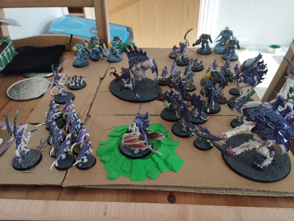 Cardboard mock up of a base/diorama with Tyranid bugs (cream and purple) with guns from Warhammer taking up most of the map. a green crater is pictured with a winged Tyranid in it with flesh coloured wings. Furthest corners away left and right of the image are space marines/wolves unfinished painted.