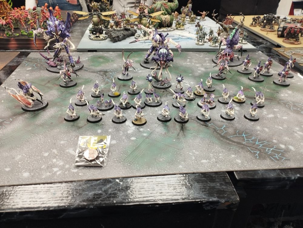 An army of Tyranids from Warhammer. Space bugs cream flesh and purple coloured armour. Smaller ones have guns in their arms. Larger ones have black and red claws. There's one with wings flesh colour like. There's a metal pin badge in a plastic clear package that says armies on parade Infront of the army.