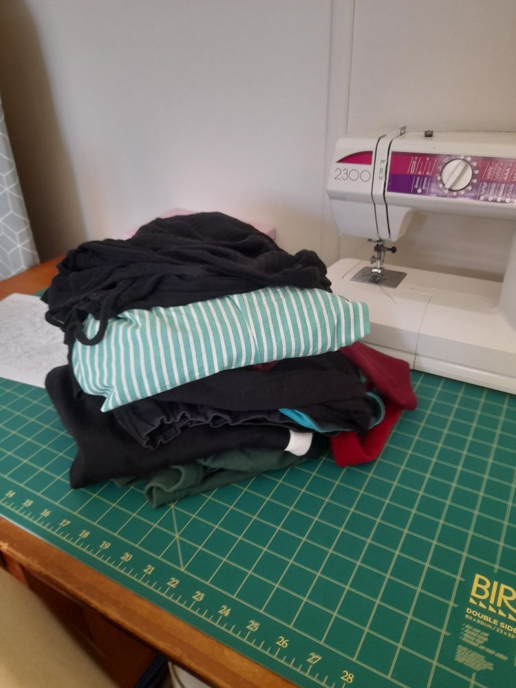 A pile of folded clothes sitting on a cutting mat on a desk. In the background is a sewing machine. 