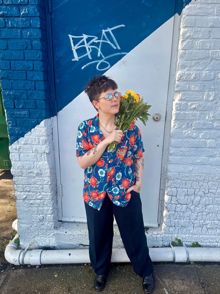 I’m wearing the blue floral Romeo button up from Romeo+Juliet, mirror sunglasses, blue slacks, and black boots. I’m holding flowers on one shoulder and looking to the side. Behind me is a door with the word BRAT in graffiti.