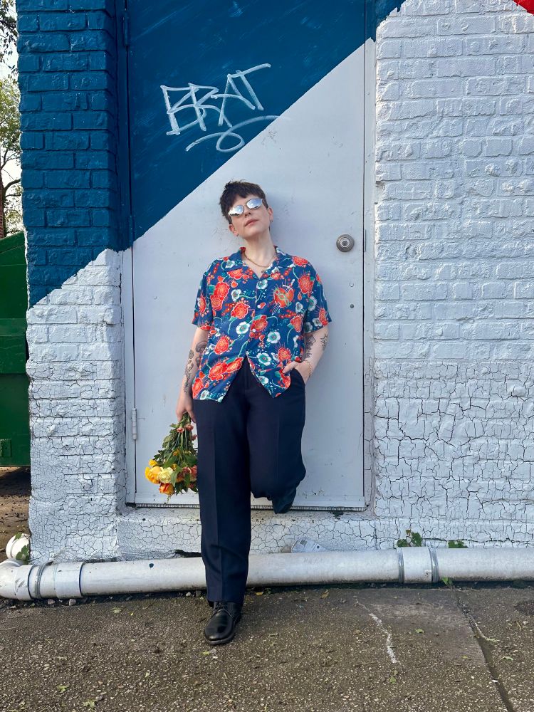 I’m wearing the blue floral Romeo button up from Romeo+Juliet, mirror sunglasses, blue slacks, and black boots. I’m holding flowers and leaning against a door with the word BRAT in graffiti on it.