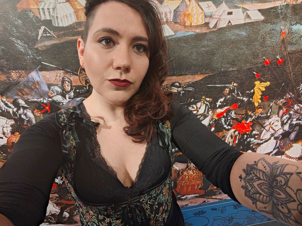 Picture of Cat Stiles, a woman in her mid thirties with dark curly hair, wearing a black top and embroidered corset, and standing in front of an illustration of the Battle of the Spurs in the Tournament Gallery at the Royal Armouries 