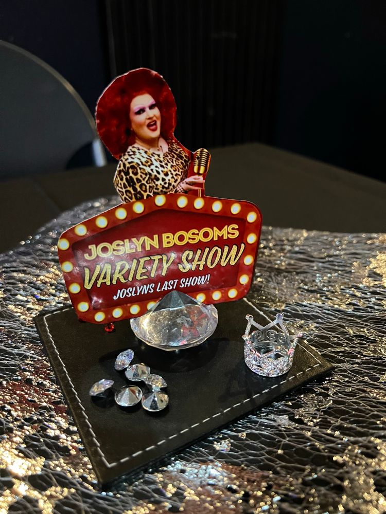 Small cardboard cutout of drag queen Joslyn Bosoms with the text “Joslyn Bosoms Variety Show. Joslyn’s last show” mounted on a plastic gemstone. 
