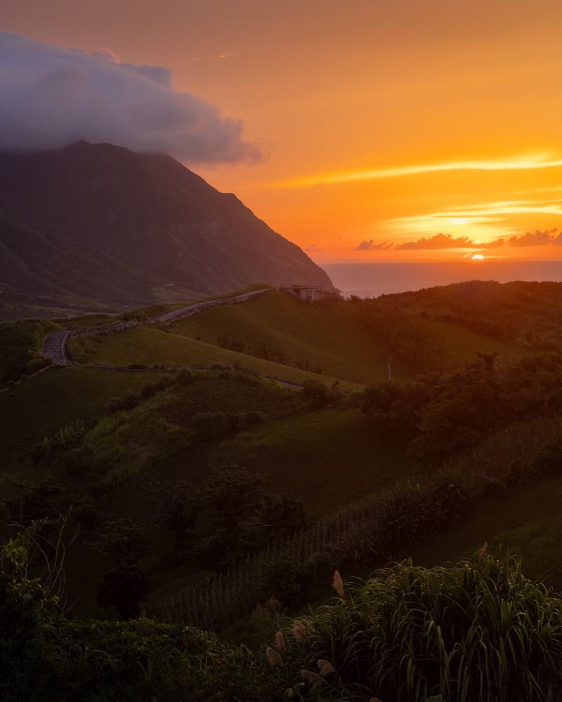 A hilly green landscape with a mountain in the background. The sun rises just above the ocean.