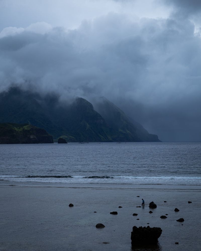Thick cloud covers a mountain at sea. A man walks on the beach