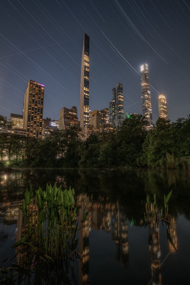 Tall buildings reflected on the Pond in Central Park. Star trails in the sky.