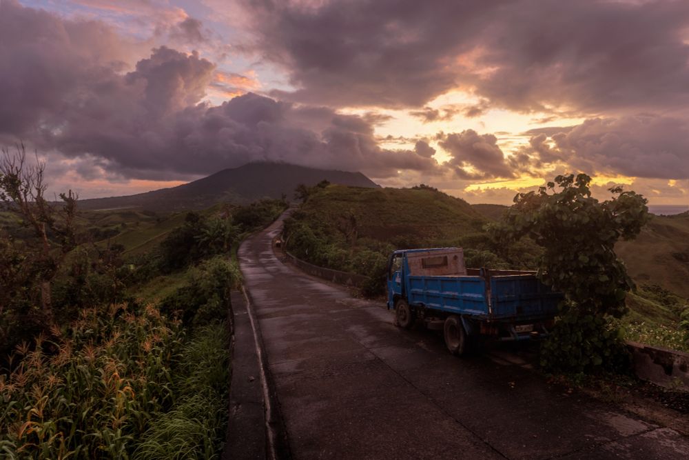 Cumulus clouds cover a mountain in the distance amidst a colorful sunrise. A road and a blue truck in the foreground