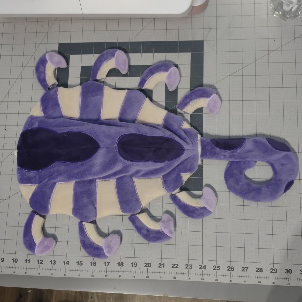 Flattened, unstuffed tail of a plush. It resembles a face hugger, with a spade center and curved tendrils protruding from the sides and end. It is purple with dark purple spots. The tips of the tendrils are light purple. Between the tendrils is beige webbing.