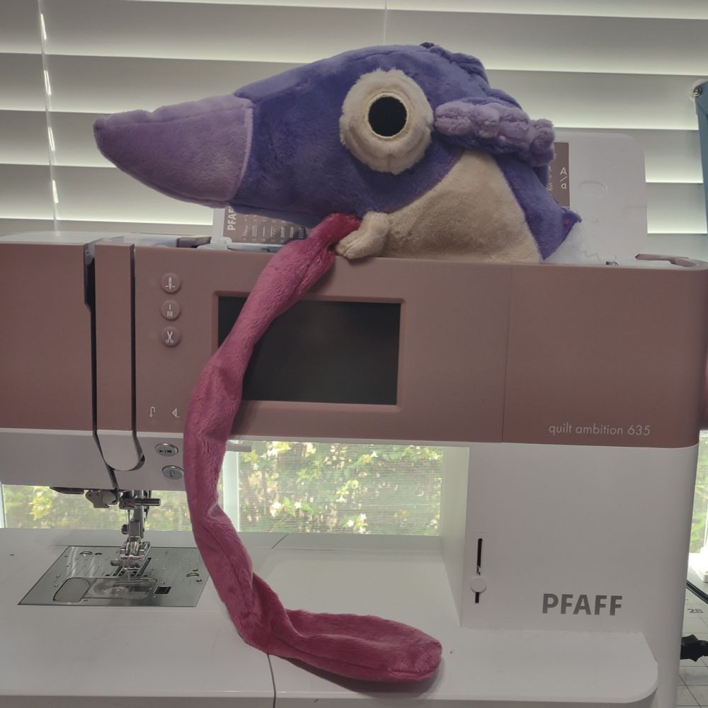 The finished plush head of a Chameleos with a long tongue. The head is sitting atop a pink and white PFAF sewing machine.