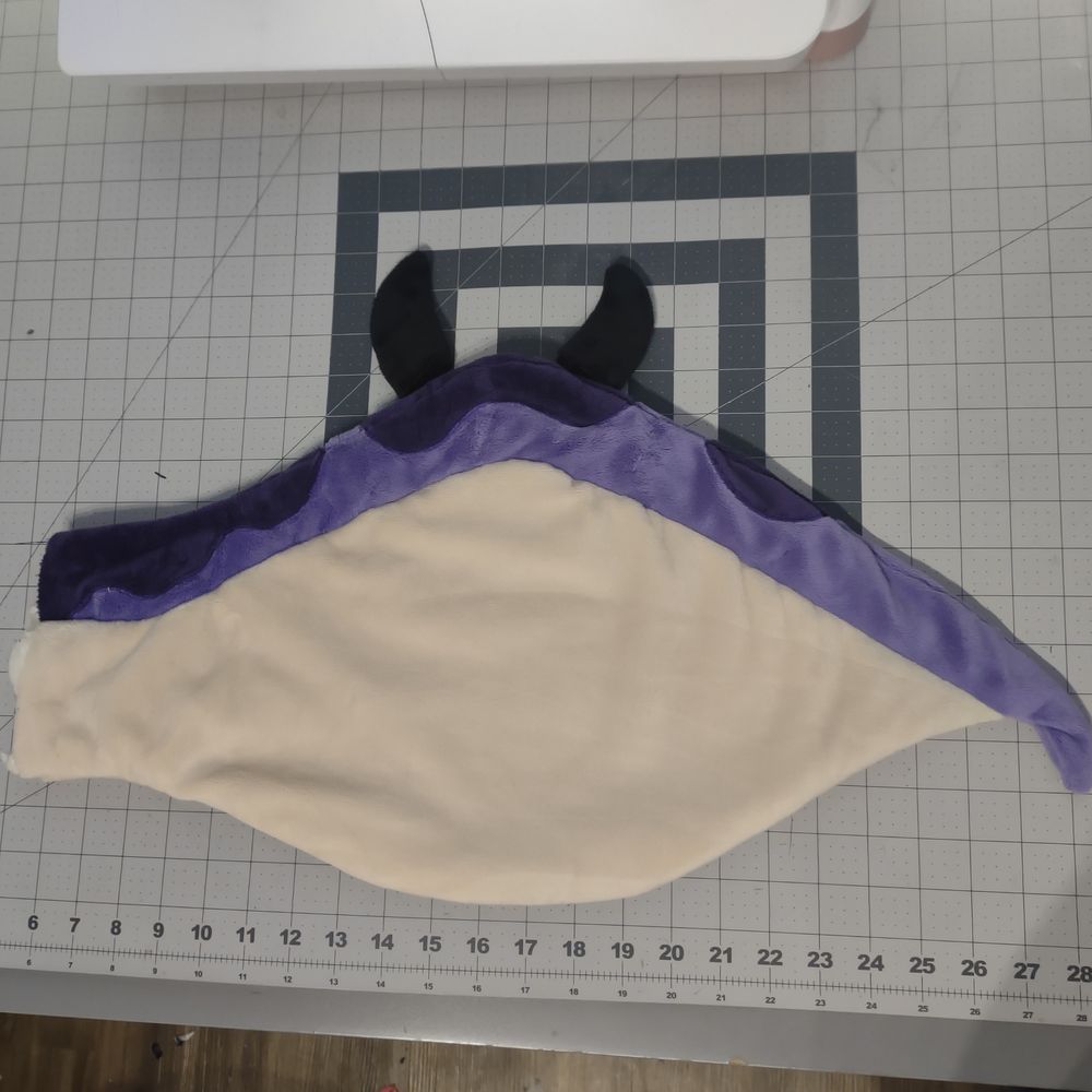 Flattened, unstuffed plush wing with black claws on the top of the wing. The wing body is beige. The arm of the wing is purple with dark purple spots.