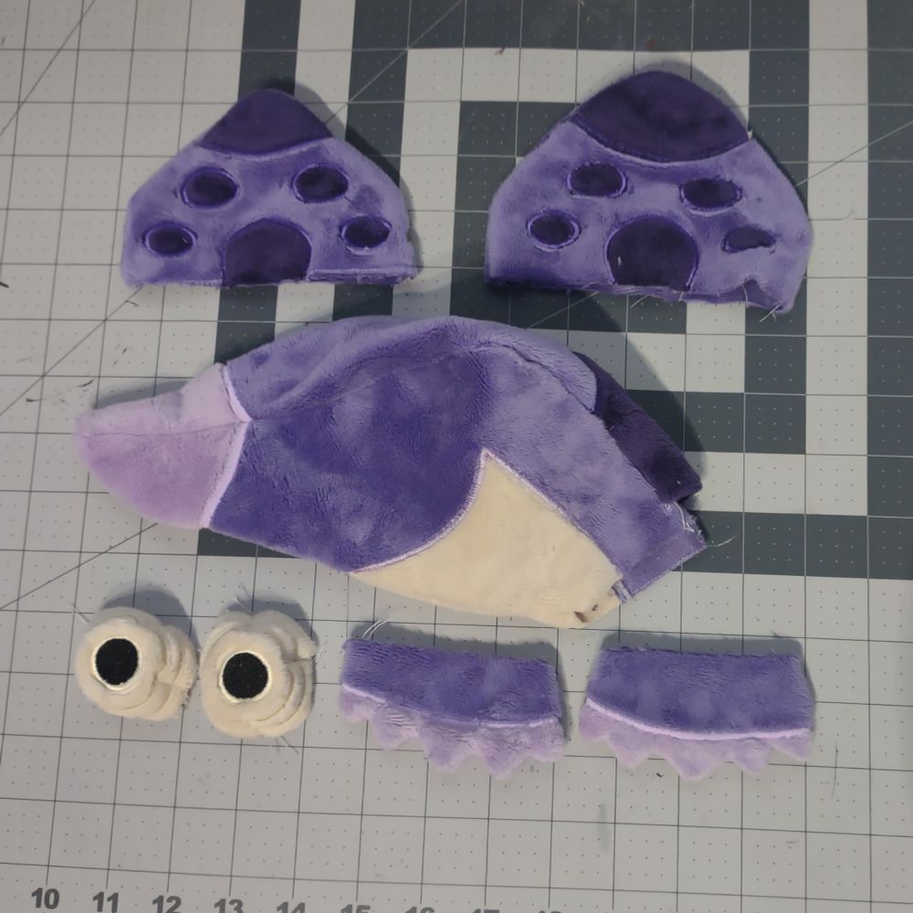Flattened, unstuffed plush pieces. Top is 2 trapezoidal purple pieces with dark purple spots. Middle is the unstuffed head: a purple conical shape with a light purple tip and beige base/throat piece. Bottom is two beige pyramids resembling chameleon eyes and two cheek ruff pieces with light purple tips.