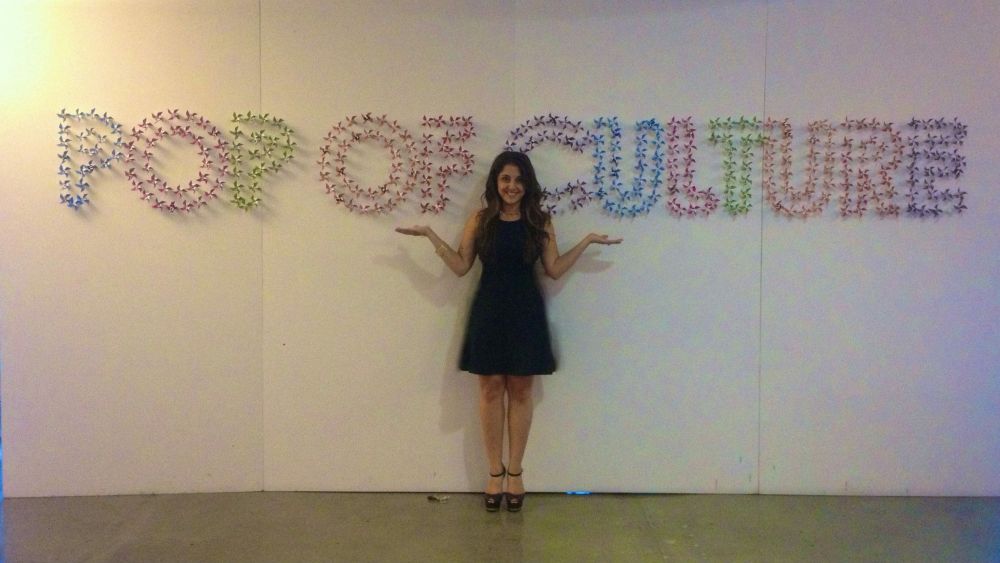 A woman in a black knee-length, sleeveless dress standing in front of a wall where the words “pop of culture” are written in bright colors. 
