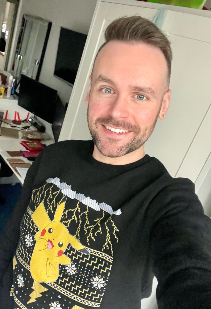 A selfie of a white male, smiling into the camera and wearing a black Christmas sweatshirt featuring Pikachu and an associated electric type motif.