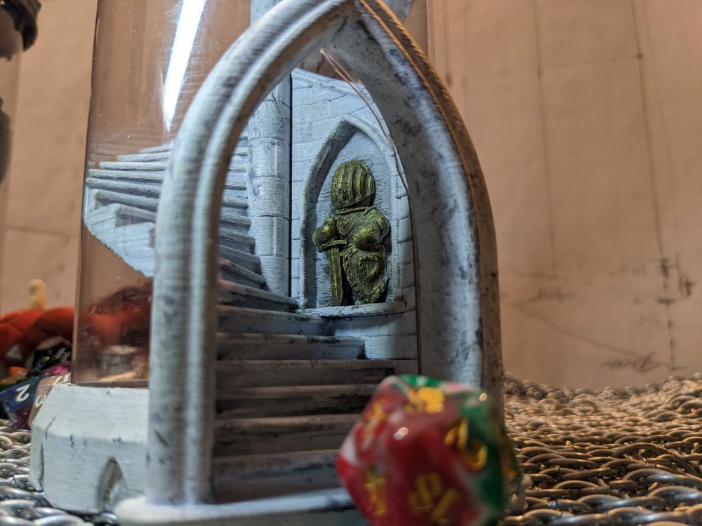 Close up of the white dice tower. A cutesy golden knight statue rests in an alcove next to the stairs.

A lucky cat coloured die rests in front of the tower partly to give scale and partly cause it's one of my favourite d20s.