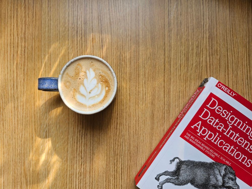 a flat white in a blue cup with passable latte art, and a copy of "designing data intensive applications" by Martin kleppmann on a wooden table.