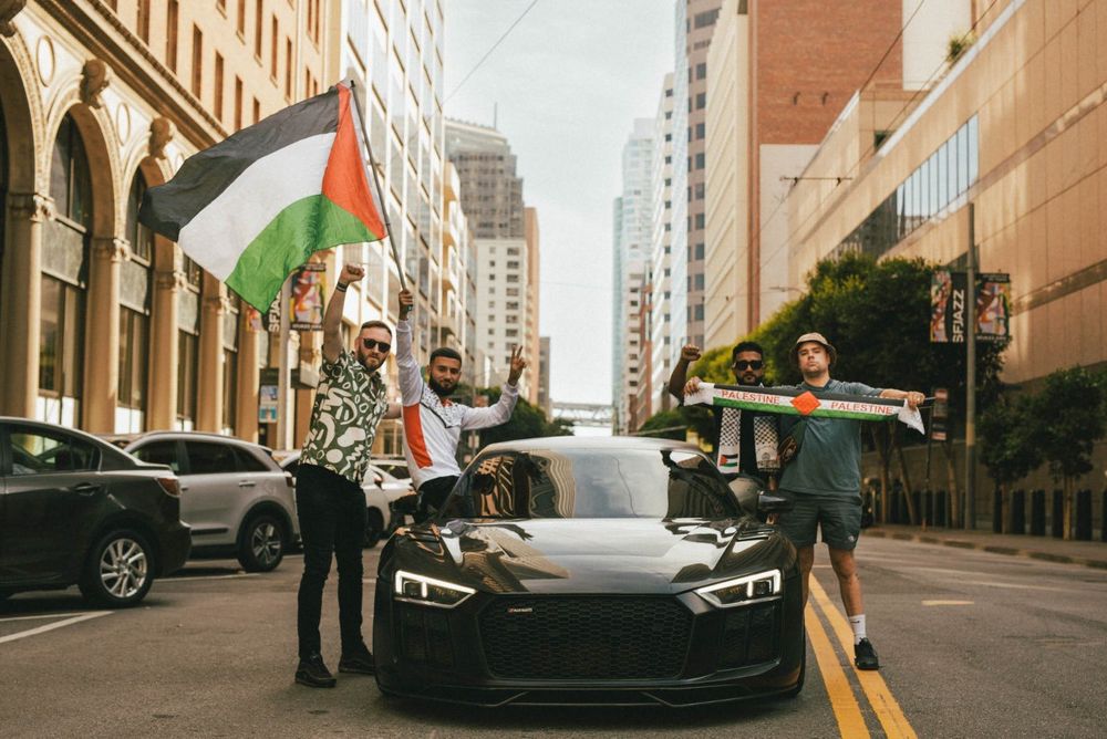 DJ Provai and Mogai Bap with two other men by a black Audi R8 holding a Palestine flag and scarf. 