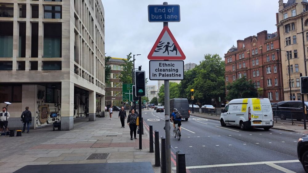 A modified "Warning: Children Crossing" street sign outside Westminster Magistrates Court. It shows children fleeing war and has the text "Ethnic cleansing in Palestine"