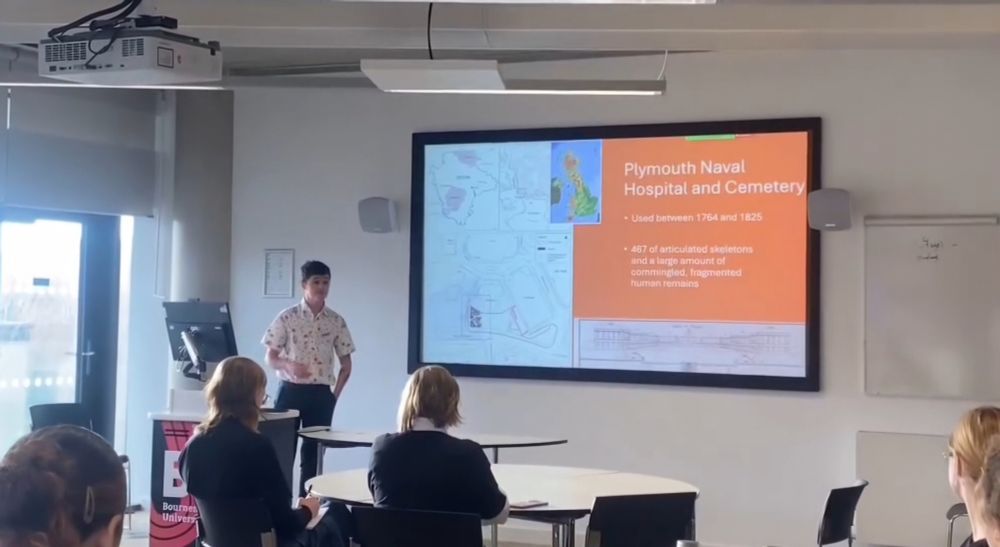 A small transgender man stood in front of a powerpoint stating "Plymouth Naval Hospital and Cemetery" 
