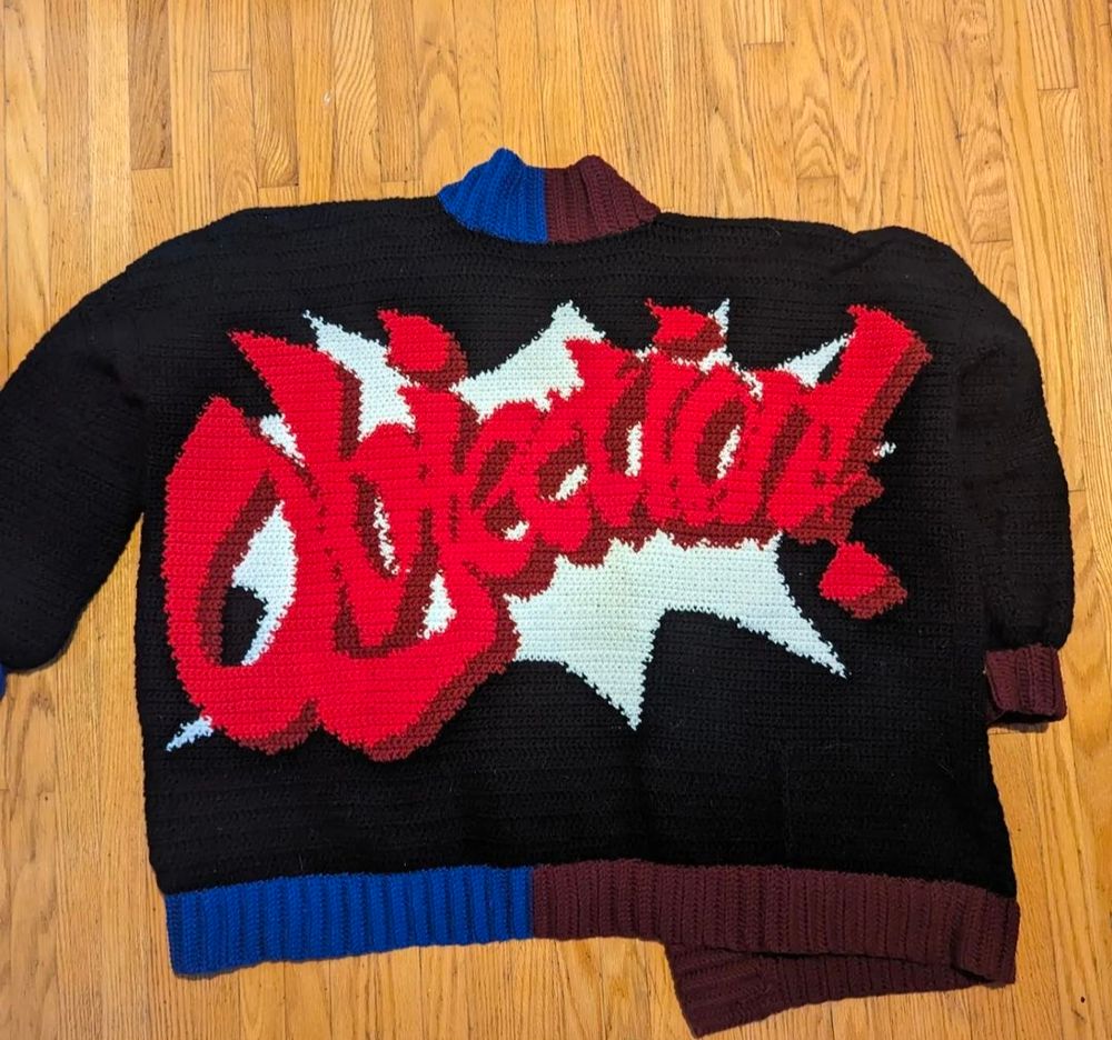 The back of a crocheted cardigan. It's black with burgundy and blue ribbing and the "objection" image from Ace Attorney across it