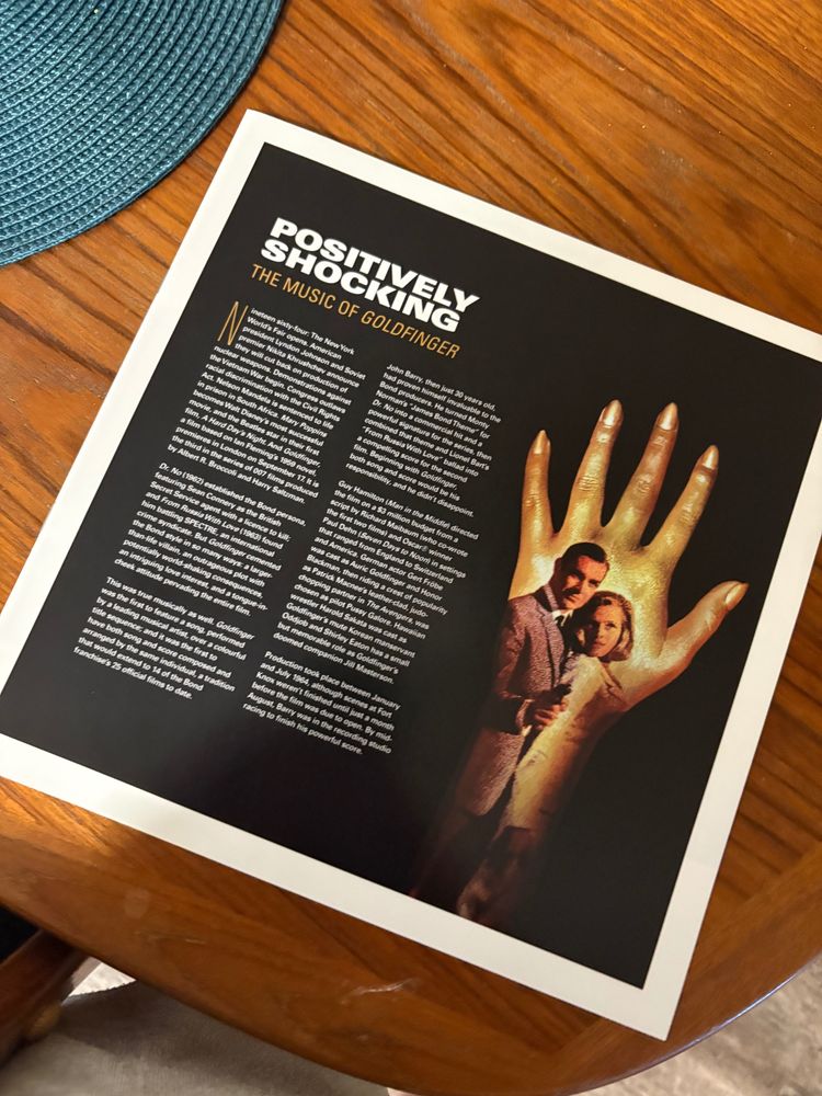 Goldfinger record insert with essay titled Positively Shocking The Music of Goldfinger. There is text too small to read and a picture of James Bond and Pussy Galore on the front.