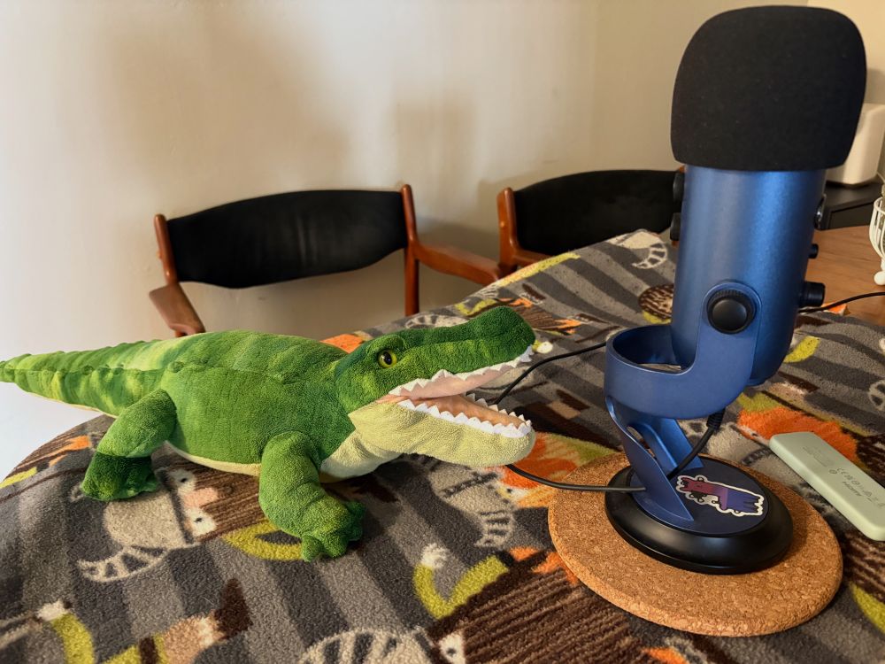 A green gator stuffy named Gator on a table facing a microphone. 