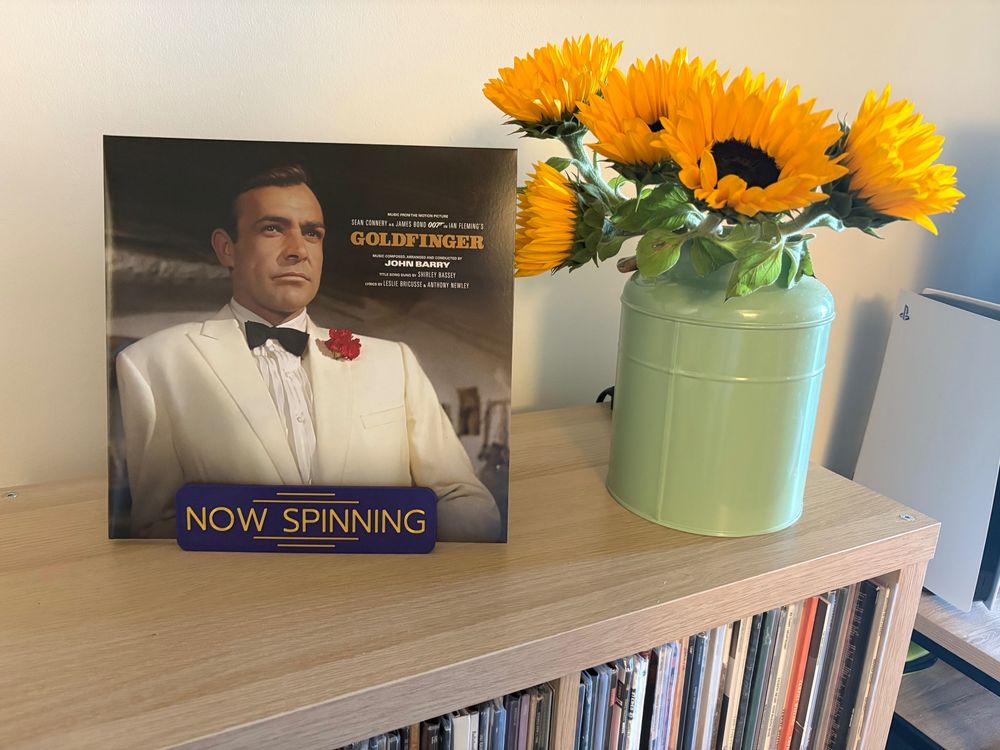 Wooden record furniture with a NOW SPINNING record shelf. The record is the score of Goldfinger, the James Bond film. Next to the record cover is a vase with sunflowers.