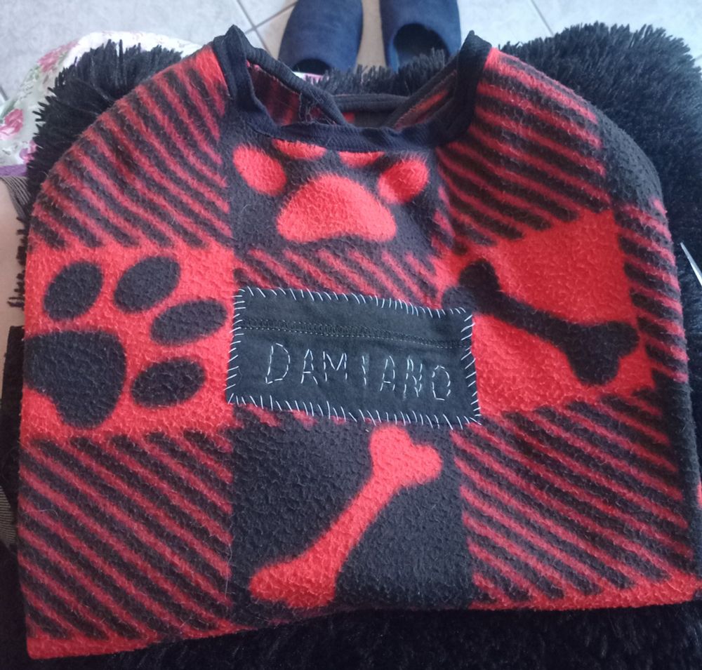 Uma roupinha pra cachorro nas cores preto e vermelho. Ela tem estampas listradas, em estilo xadrez intercaladas com ossinhos e patinhas. Tem um patch preto com o bordado branco com o nome Damiano nele.