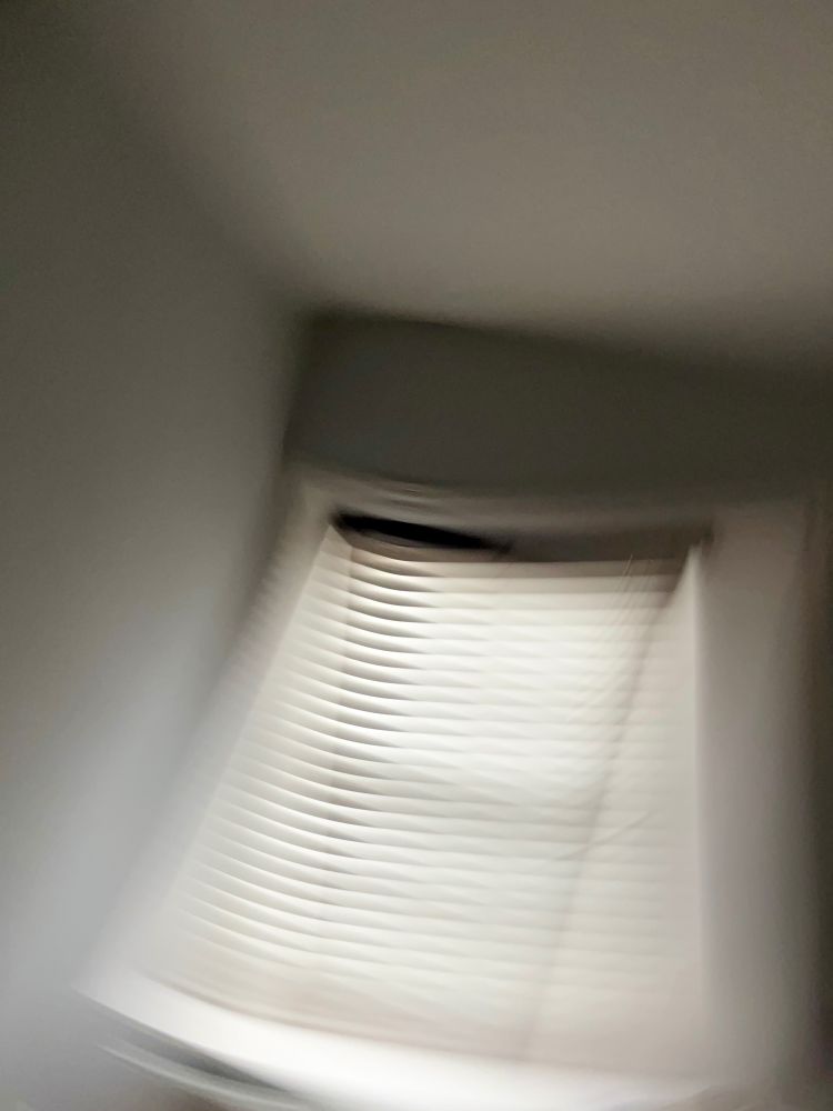 Shuttered window. Abstract. 
The photo shows a blurred view of a window with blinds, captured in a room with light blue walls.  I’ve captured movement deliberately. The blinds are partially open, allowing some light to filter through, creating a contrast between light and shadow. It’s 9:32am