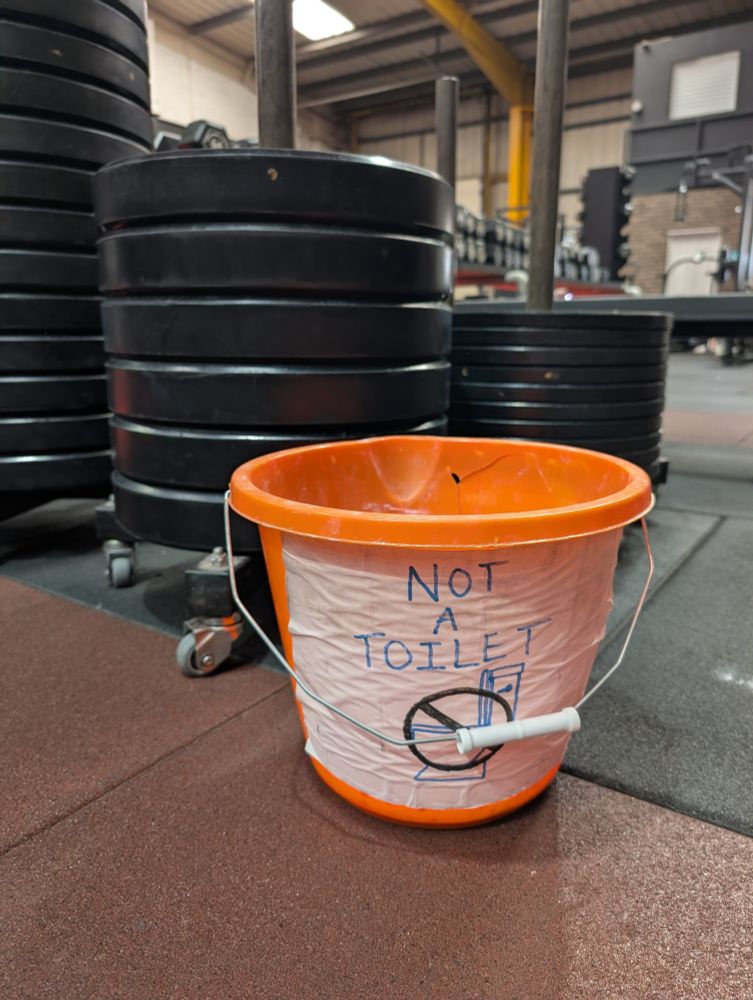 An orange bucket of chalk sits in front of barbell plates. A hand written sign on the bucket states "NOT A TOILET"