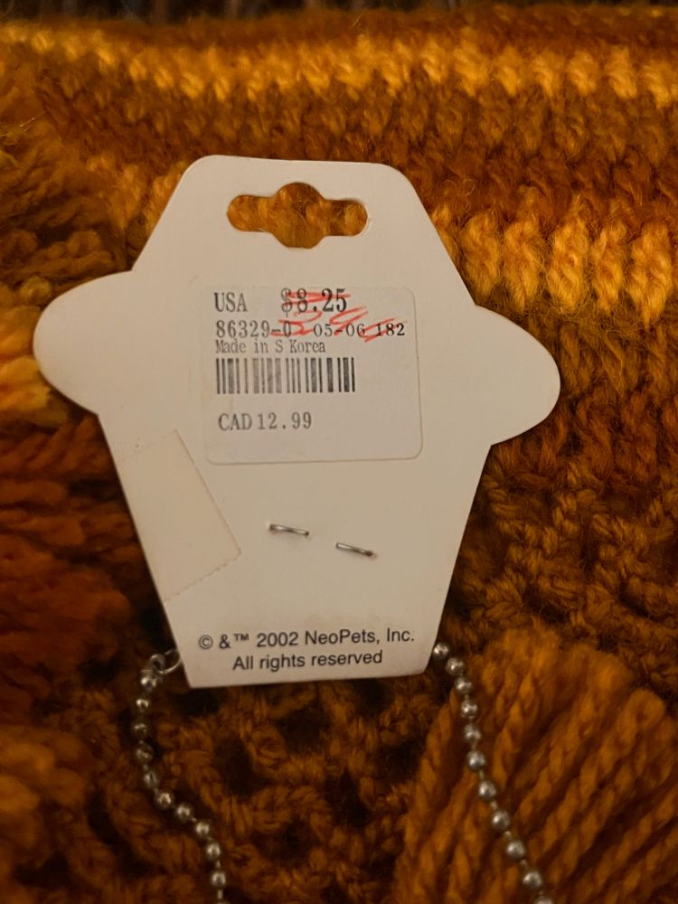 back of the tag of the neopets necklace still resting on the yellow orange crocheted blanket. USD $8.25 had been crossed out in red and written in the same color it says $3.90. Below that is a string of numbers, made in S. Korea, a barcode, and CAD 12.99. At the bottom of the tag it reads, copyright (in circle symbol form) & TM 2002 NeoPets, Inc. All rights reserved.