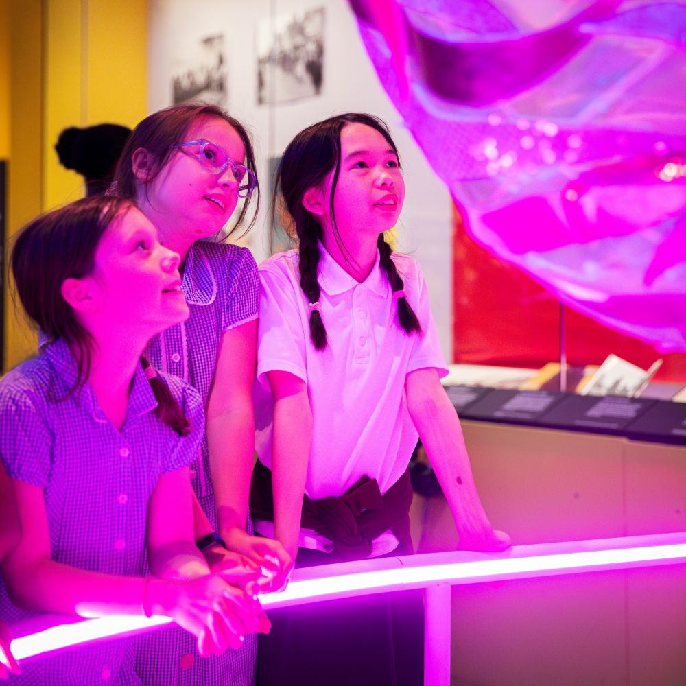 Three primary school aged children are in an exhibition space. They're looking up in wonder at a large glowing, pink ball.
