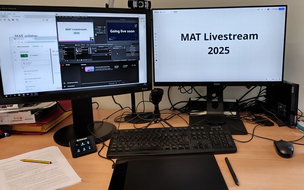 Two computer screens, and a mess of tech peripherals like a tablet and microphone and a mini stream deck. One screen says MAT Livestream 2025 in large letters.