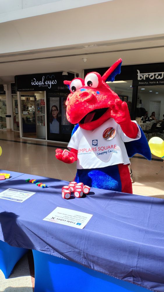 A large red dragon mascot (wearing a T-shirt identifying him as the mascot for Templars Square shopping centre) attempts a maths activity with dice at the Oxford Maths Festival.