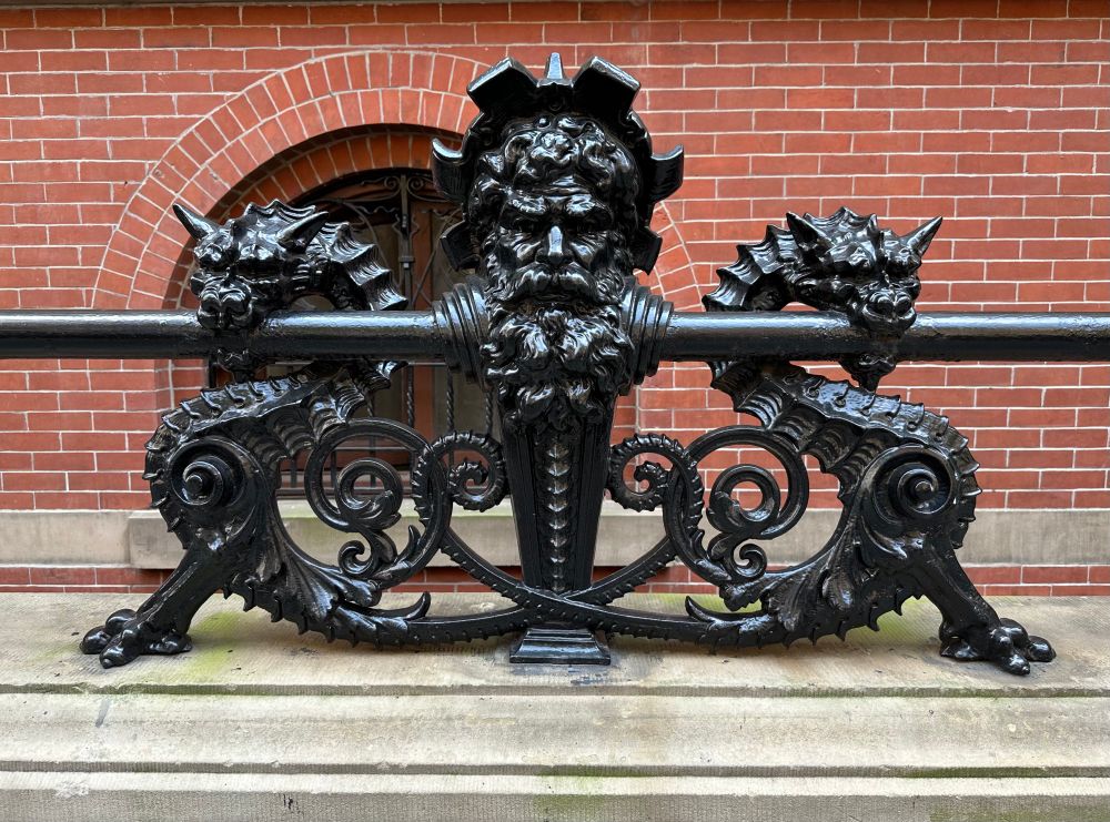 red brick background with an elaborate black cast iron railing decorated with a bearded man’s head bookended by two dragons 