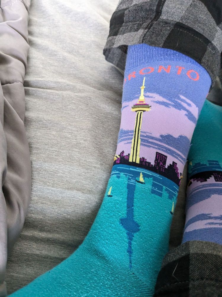 Socks depicting the Toronto skyline. The waters of Lake Ontario are dotted with sailboats and the reflections of buildings, primary of which is the CN Tower. All the buildings can be seen atop a backdrop of the morning sky beneath the word "Toronto"