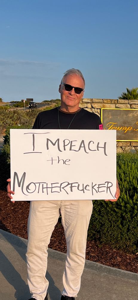 A man in a black T-shirt and light pants, holding a sign that says impeach the motherfucker