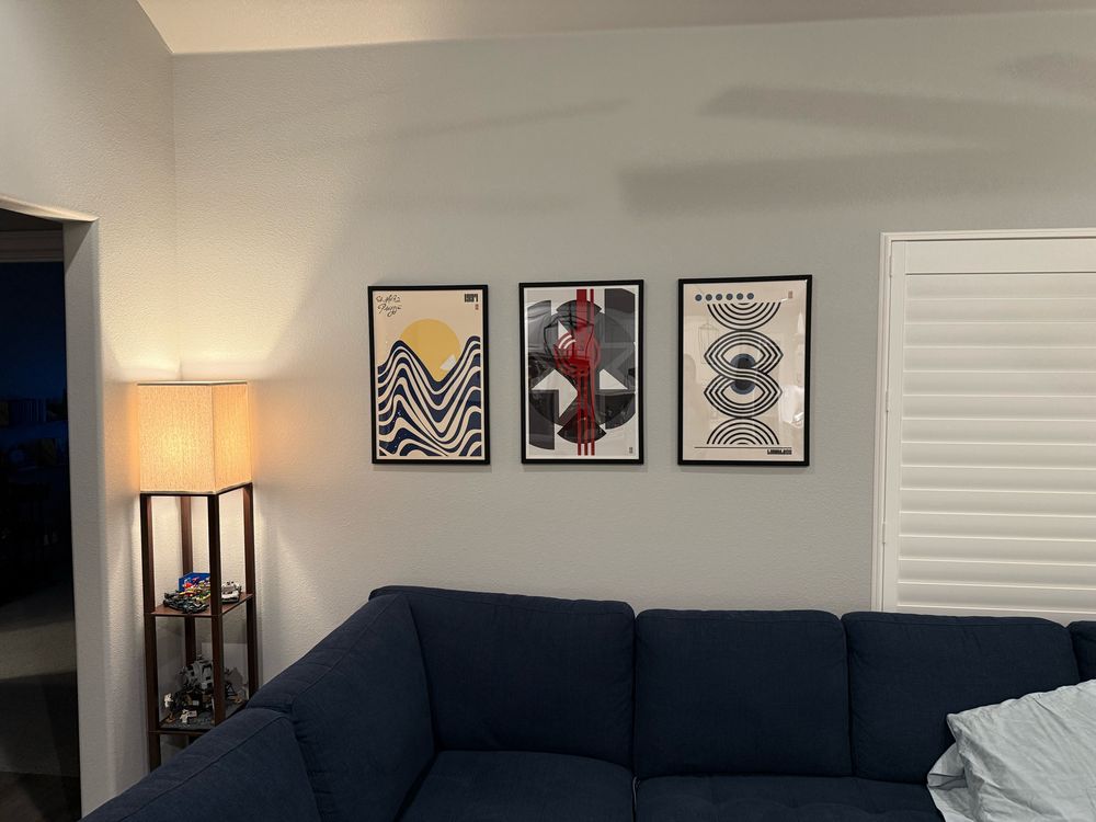 Three prints hanging on a wall side by side above a couch. 
Left most print shows abstract blue waves with a small boat silhouette and a yellow circle in the background. 

Center print is a handful of geometric shapes in darker grey making outlines of a star with red lines going through the middle. 

Right most is a handful of abstract lines in semi circles and ovals with 6 dots across the top. 